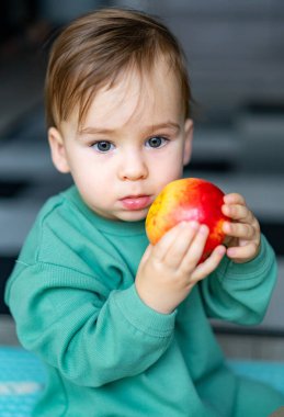 Yerde otururken parlak bir elma tutan küçük bir çocuk. Mavi gözlü küçük bir çocuk kırmızı elmanın tadını çıkarıyor.