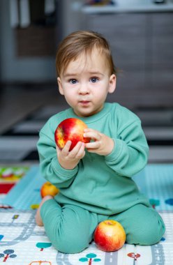 İçerideki renkli bir paspasın üzerinde oturan çocuk elma tutuyor. Yeşil elbiseli bir çocuk oyun hasırının üzerinde oturur.