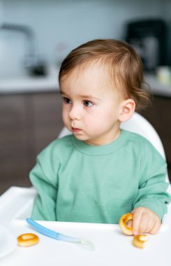 Bebek gün boyu yüksek sandalyede abur cuburları keşfediyor. Yeşil elbiseli genç bir çocuk yüksek bir sandalyede oturur ve tuhaf bir şekilde parlak bir mutfakta atıştırmalıkları inceler..