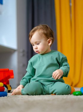 Bebek, rahat bir oyun odasında yumuşak bir halıda renkli oyuncakları keşfediyor. Genç bir çocuk halının üzerinde oturuyor, neşeli bir atmosferde parlak oyuncaklarla meşgul..