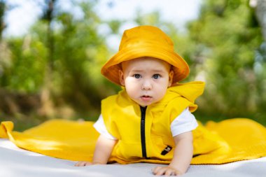 Sarı kıyafetli bir bebek parkta güneşli bir günün tadını çıkarıyor. Bir bebek sarılı bir elbise ve şapkayla, yeşil bir battaniyenin üzerinde emekler.