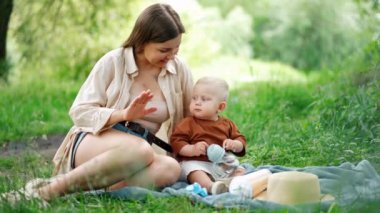 Anne ve küçük oğlu yazın çimlerin üzerinde otururlar. Annem el çırpıyor ve şarkı söylüyor ama bebek şişeden su içiyor..