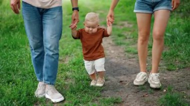 Tanınmayan anne ve baba, bebeklerini açık havada ellerinde gezdiriyorlar. Şirin çocuk yazın parkta ilk adımlarını atıyor..