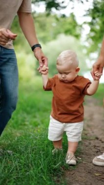 Tanınmayan ebeveynler bebeklerini elleriyle tutarlar. Anne ve baba dans ediyor ama çocuk onları hareketsiz izliyor. Bulanık doğa arkaplanı. Dikey video.