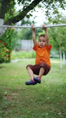 Turuncu tişört giyen ve kahverengi şortlu küçük çocuk yatay çubukta asılı duruyor. Çocuk ileri geri sallanıyor ve ayaklarını kaldırıyor. Dikey video.