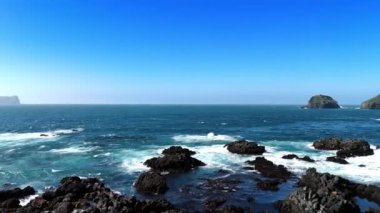 Kıyıda sudan çıkmış siyah kaya parçası. Beyaz dalgalar kayalara çarpıyor. Dik uçurum, su manzarası üzerinde yükseliyor..