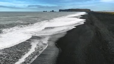 Deniz dalgaları sahile siyah kumla vuruyor. Arka planda sarp kayaların siluetleri. İzlanda vahşi doğası.