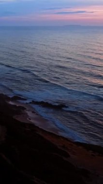 Küçük dalgalar kayalık kıyıya çıkar. Batan güneş denizin dalgalı yüzeyine yansıyor. Hava görüntüsü. Dikey video.