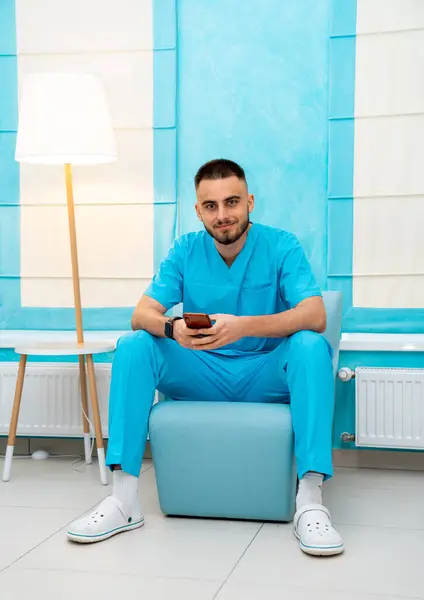 stock image Doctor in blue uniform at clinic. A healthcare worker in a blue uniform is seated comfortably, looking at a mobile phone in a modern clinic setting.