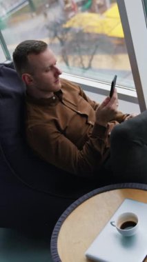 İş adamı ofis penceresinden rahatlıyor. Elinde telefon olan bir adam rahat bir kanepenin camından şehre bakıyor. Yaşam dengesi kavramı üzerinde çalış. Üst Manzara. Dikey video