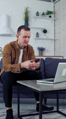 Evden çalışan genç bir erkek. Evde dizüstü bilgisayarda çalışan serbest çalışan bir adamın portresi kanepede oturuyor. Karantina uzaktan kumandası. Dikey video
