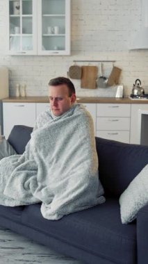 Ateşi olan bir adam. Evdeki kanepede otururken kendini üşümüş ve sıcak bir battaniyeyle örtülmüş hisseden genç bir adam. Covid salgını sırasında battaniyeye sarılı hasta adam. Dikey video