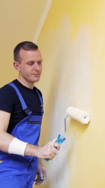Oda makyajı. Duvarı boyayan genç bir adamın portresi. Duvarı yeni bir renge boyayan bir işçi. Kapalı alandaki bir merdivenden çıkan boya silindiriyle. Dikey video