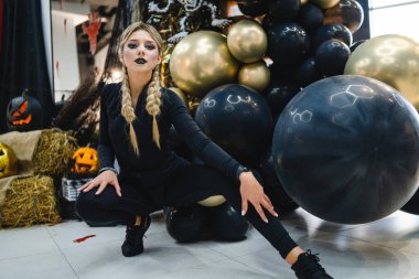 Cadılar Bayramı dekoru arasında siyahlı genç bir kadın. Örgülü genç bir kadın, Cadılar Bayramı temalı süslemeler ve balonlarla çevrili siyah kıyafetler içinde dramatik bir şekilde poz veriyor..