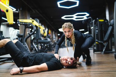 Dramatik bir dönüşle jimnastik salonu. Bir fitness eğitmeni spor salonunda bir adamın yanında diz çöker ve modern bir spor salonunda eşsiz bir eğitim metodu gösterir..