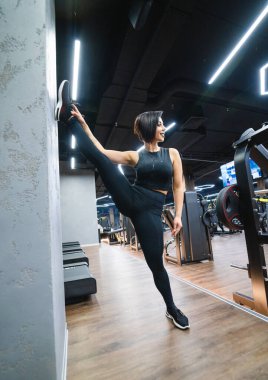 Modern bir spor salonunda esneme hareketleri yapan bir kadın. Aktif giyimli bir kadın, çağdaş bir spor salonunun duvarına karşı bacak esneme hareketi yapıyor.