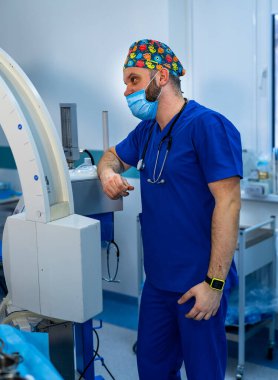 Ameliyat sırasında mavi önlüklü ve şapkalı bir cerrah. Mavi önlüklü ve renkli şapkalı bir cerrah tıbbi bir cihazın yanında duruyor. Parlak bir hastane odasında ameliyat için hazırlanıyor..