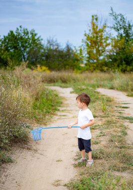 Çimenli bir alanda ağla doğayı keşfeden küçük bir çocuk. Beyaz tişörtlü ve şortlu bir çocuk çimenli bir yolda geziniyor.