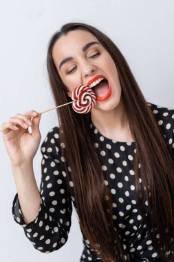 Genç bir kadın parlak kırmızı rujlu bir lolipopun tadını çıkarıyor. Kadın neşeli bir şekilde çizgili bir lolipopu ısırıyor, canlı kırmızı rujunu ve polka noktalı kıyafetiyle neşeli ruhunu gösteriyor..