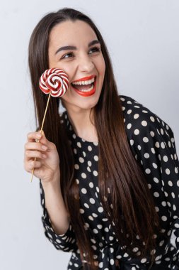 Puantiyeli elbiseyle lolipopun keyfini çıkaran neşeli bir kadın. Uzun saçlı bir kadın neşeyle sarmal bir lolipop tutar.