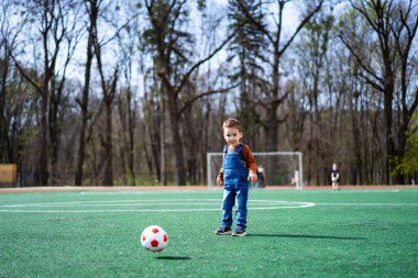 Parkta güneşli bir günde futbol oynayan küçük bir çocuk. Parlak, güneşli bir günde ağaçlarla çevrili yeşil bir arazide tulumlu bir çocuk futbol topunu tekmeliyor..