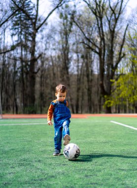 Parkta güneşli bir günde futbol oynayan küçük bir çocuk. Tulumlu bir çocuk açık mavi gökyüzünün altındaki ağaçlarla çevrili çimenli bir arazide futbol topunu tekmelemekten hoşlanır..
