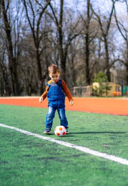 Parkta güneşli bir günde futbol oynayan küçük bir çocuk. Parkta güneşli bir günde tulumlu bir çocuk futbol oynuyor.