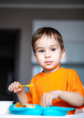 Küçük çocuk öğleden sonra evde yemeğin tadını çıkarıyor. Turuncu tişörtlü bir çocuk bir masada oturur ve öğle vakti mavi bir kaşıkla mutlu bir şekilde yemek yer..