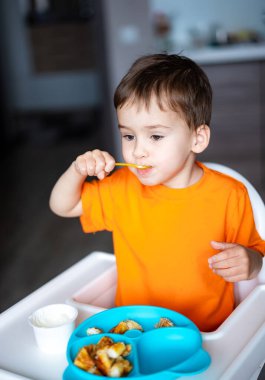 Genç çocuk modern bir mutfakta renkli bir yemeğin tadını çıkarıyor. Turuncu tişörtlü bir çocuk, açık mutfak ortamında mavi bir tabaktan mutlu bir şekilde yemek yer..
