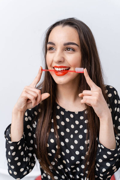 Young woman playfully poses with toothbrush and red lipstick. A young woman smiles cheerfully, holding a toothbrush with bright red lipstick while playfully posing against a light background.