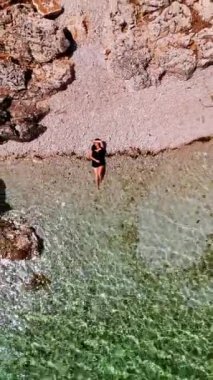 Kadın kayalık plajda güneşleniyor. Arnavutluk sahilinde dinlenin. İnsansız hava aracı temiz suyun üzerinde yükseliyor. Dikey video.