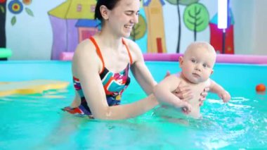 Renkli mayolu gülümseyen kadın bebeğe yüzmeyi öğretir. Ciddi sarışın çocuk sakin kalır. Kapat..