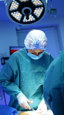 Mavi üniformalı bir cerrah forseps ve iplik kullanır. Doktor, kesiği operasyonun son aşamasında dikiyor. Dikey video.