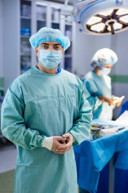 Mavi ameliyat elbisesi giymiş bir adam ameliyat masasının önünde duruyor. Mavi bir şapka ve mavi bir maske takıyor. Arkasında başka biri var.