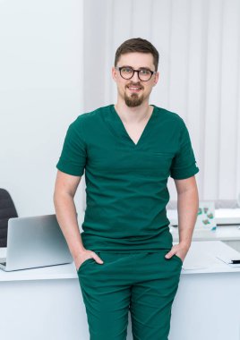 Yeşil önlüklü bir adam üstünde laptop olan bir masanın önünde duruyor. Gülümsüyor ve mutlu.