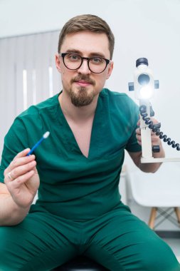 Yeşil laboratuvar önlüklü bir adam elinde kalem ve ışıklı bir kamera tutuyor. Gülümsüyor ve bir doktor.