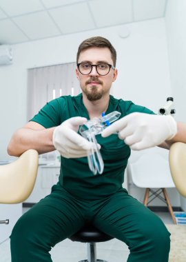 Yeşil laboratuvar önlüklü bir adam tıbbi bir alet tutuyor. O bir gözlük takıyor ve bir taburede oturuyor. Olay yeri tıbbi bir ortamda, muhtemelen dişçi muayenehanesinde.