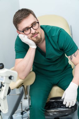 Yeşil önlüklü bir adam elinde başıyla bir sandalyede oturuyor. Sol elinde beyaz bir eldiven var.