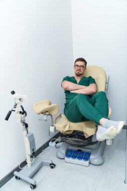 Yeşil önlük giymiş bir adam tıbbi sandalyeye oturur. Sandalye görünmeyen bir makineye bağlı.