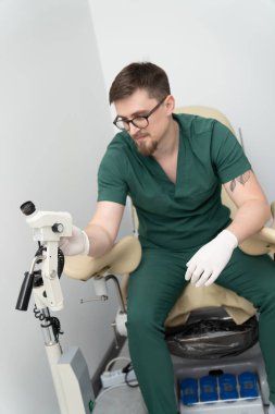 Yeşil laboratuvar önlüklü bir adam elinde tıbbi bir aletle bir sandalyede oturuyor.