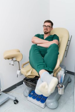 Yeşil önlük giymiş bir adam hastane yatağında oturuyor. Gözlük takıyor ve rahat bir durumda.