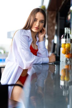 Beyaz gömlekli ve kırmızı bikinili bir kadın bir barda oturuyor. Kameraya bakıyor ve rahatlamış durumda. Önündeki masada birkaç portakal var ve bir şişe de görünür durumda.