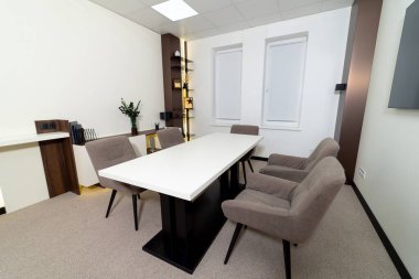 Büyük bir masa ve altı sandalyeli bir konferans odası. Sandalyeler kahverengi ve masa beyaz. Masanın üzerinde saksı bitkisi ve masanın üzerinde vazo var. Odanın bir profesyoneli var.