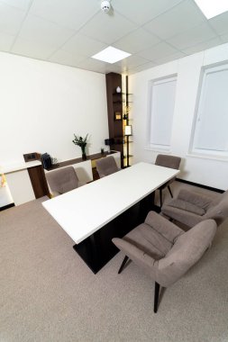 Masası ve sandalyeleri olan küçük bir ofis. Masa beyaz ve sandalyeler kahverengi. Masanın üzerinde saksı bitkisi var.