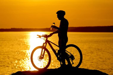 Bir adam su kenarında bir kayanın üzerinde bisikletle duruyor. Kask takıyor ve su şişesinden su içiyor. Arka planda güneş batarken sahne huzurlu ve huzurlu.