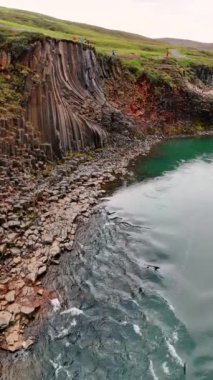 Nehrin üzerinden uçup dağların arasından sütun kayalarıyla akacağız. Turistler İzlanda 'da kayalık arazinin yanından geçerler. Dikey video.