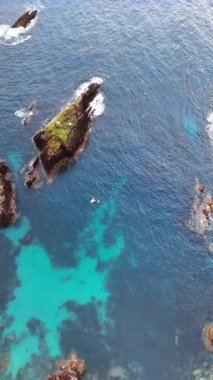 Mavi sulardan sarkan ayrı kayalar. Suda bir turkuaz nokta kıyıya yakın görülebilir. Dikey video.