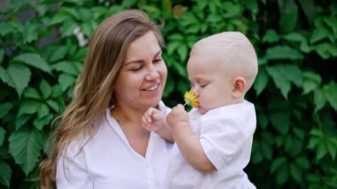 Gülümseyen uzun saçlı kadın kucağında küçük sarışın bir oğlan çocuğu tutuyor. Küçük çocuk elinde bir çiçek tutuyor ve koklaması için annesine veriyor..