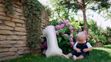 Yeşil çimenlerin üzerinde oturan sevimli bebek çiçek yapraklarıyla oynuyor. Çocuğun yanında bir numaralı balon var. İlk doğum günü konsepti.