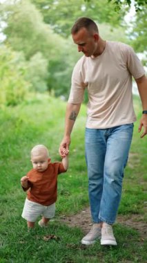 Tişört giyen ve kot pantolon giyen bir adam bebeğiyle yürüyor. Çocuk yürümeyi öğreniyor, tökezliyor, düşüyor ve babası onu kaldırıyor. Dikey video.